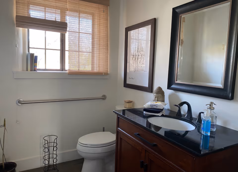Small bathroom with a toilet, wooden vanity with black countertop and sink, mirror and framed art, a window with a bamboo blind, and a wall grab bar.