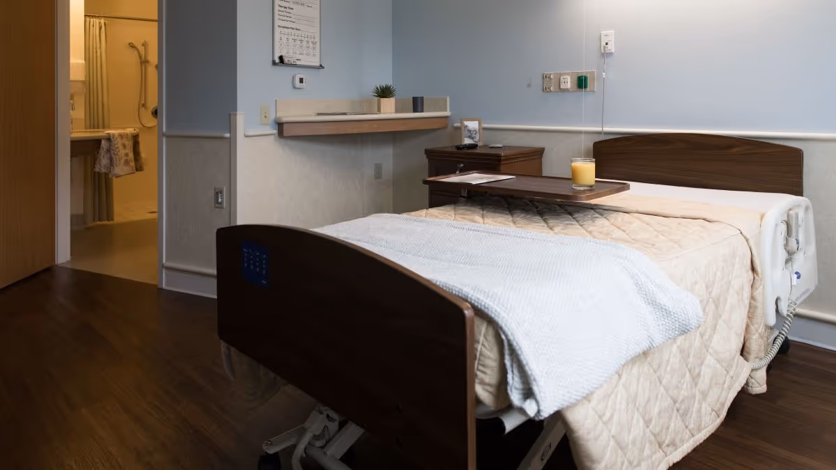 A neatly made hospital-style bed with a beige quilt and a light blue blanket in a room. A wooden bedside table with a small plant and a framed photo is next to the bed. A tray with a glass of orange juice is placed on the bed. The room has light blue walls and wood flooring. An open door reveals a bathroom with a shower and grab bars.