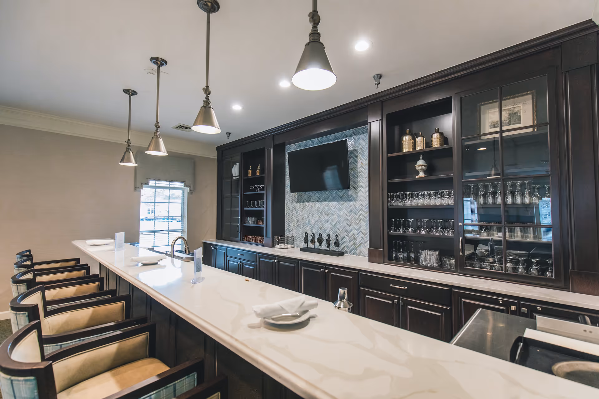 Interior view of a modern bar area with a long white marble countertop, several high chairs with armrests, dark wooden cabinetry with glass shelves displaying glassware, a wall-mounted TV, and pendant lights hanging from the ceiling.