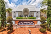 Front entrance of The Veranda Club building with its sign, manicured shrubs, and white facade under a blue sky.