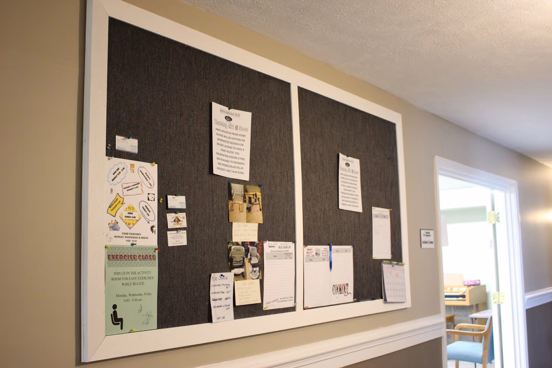 Large bulletin board displaying flyers, photos, and notices mounted on a hallway wall next to an open doorway.