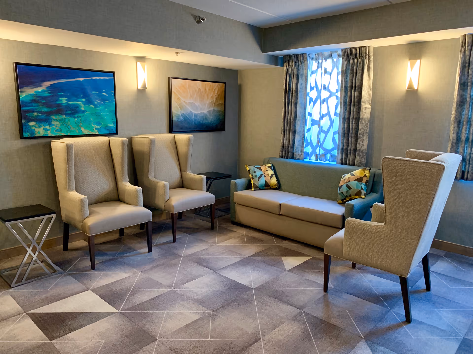 A cozy seating area in a supportive living facility featuring a green sofa with two patterned pillows, three beige wingback chairs, two small black side tables, two framed abstract paintings on the wall, and a window with patterned curtains. The floor has a geometric patterned carpet and the walls are painted in a soft neutral tone with wall-mounted lights.