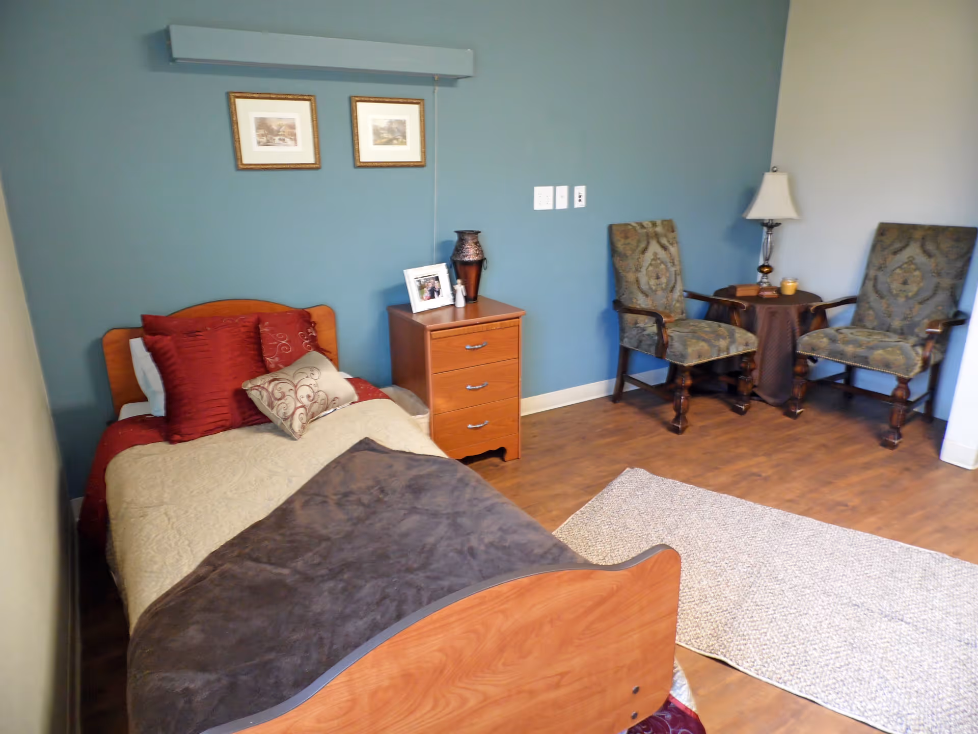 A cozy nursing facility bedroom with a single bed made with beige and burgundy bedding, a wooden nightstand with a photo frame and decorative vase, two upholstered armchairs with a small round table and lamp between them, and two framed pictures hanging on a blue accent wall.