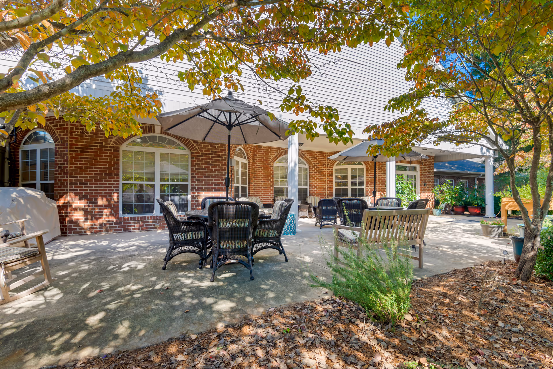 Outdoor patio area at Brookdale Robinwood with several black wicker chairs and tables under large umbrellas. The patio is adjacent to a brick building with large windows and white columns. Trees with green and orange leaves provide partial shade, and there are various potted plants and garden beds around the patio.