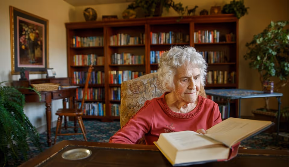 An elderly woman with white curly hair wearing a red top is sitting in a cozy room reading a large book. Behind her is a wooden bookshelf filled with books, a wooden chair, a small table with a basket, a framed floral painting on the wall, and some green plants.