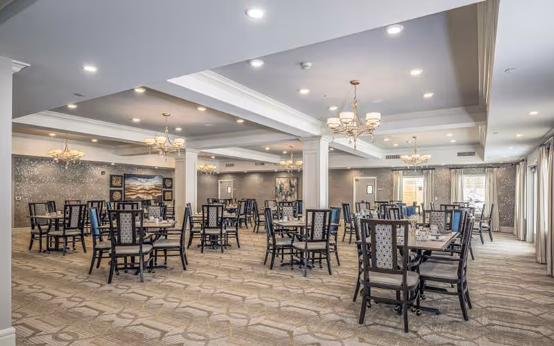 Spacious dining room with multiple tables and chairs arranged neatly. The room features chandeliers hanging from the ceiling, large windows with curtains allowing natural light, and decorative wall art. The carpet has a geometric pattern and the overall ambiance is bright and welcoming.