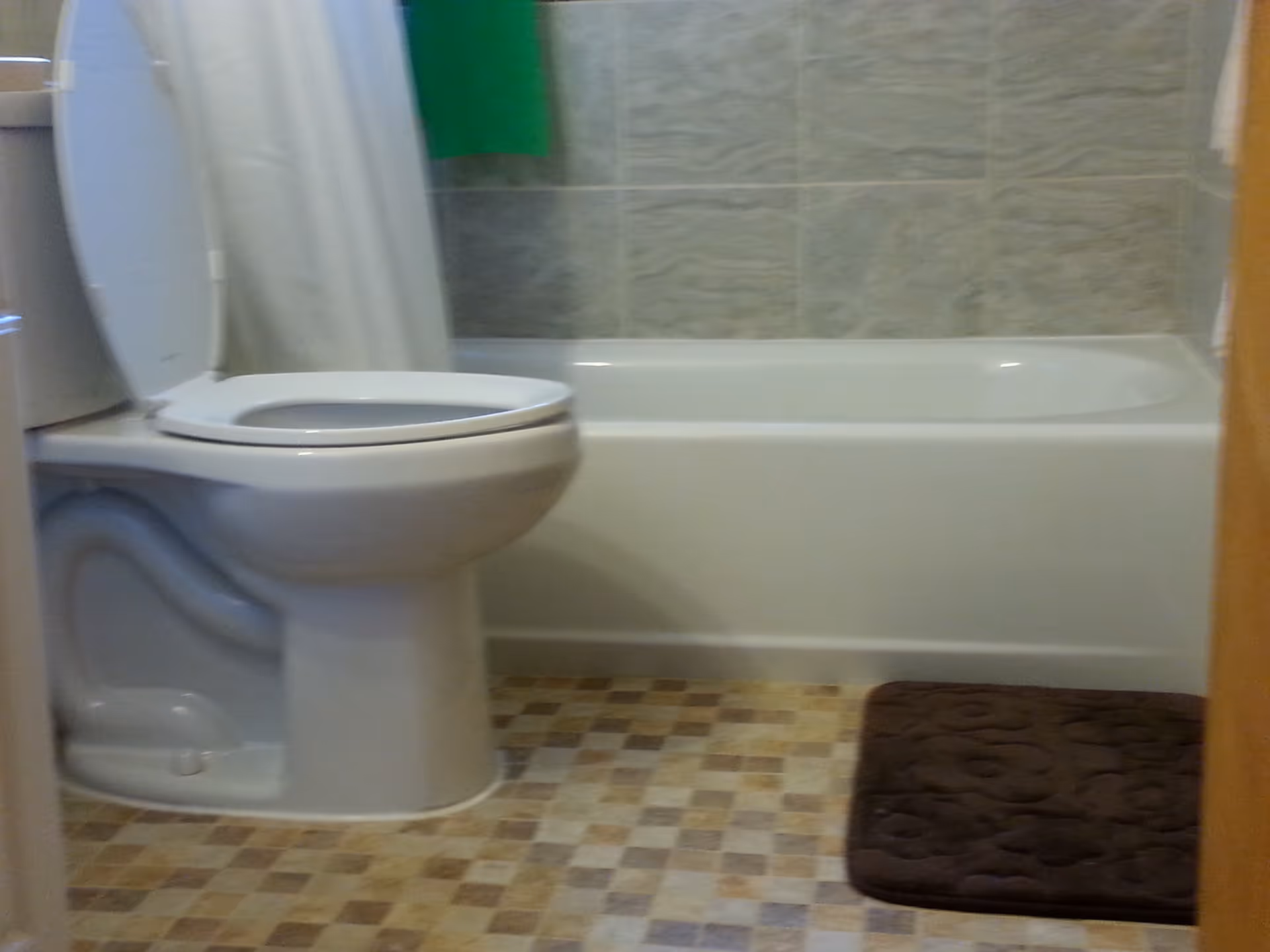 A bathroom with a white toilet with the lid open, a bathtub with a white shower curtain, tiled walls, and a brown bath mat on a checkered floor.