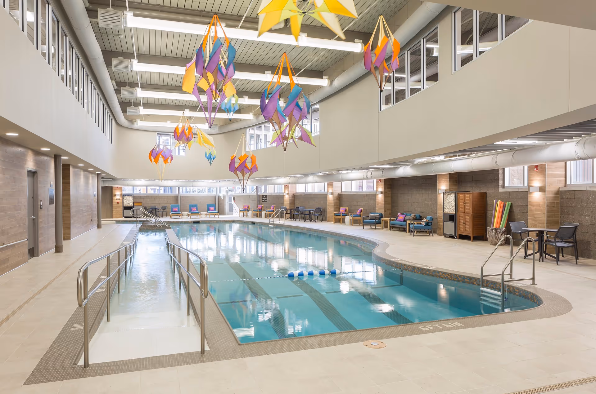 Indoor swimming pool area with a ramp and stairs for access, surrounded by seating including chairs and lounge chairs with colorful cushions. The ceiling is high with large windows and colorful hanging decorations shaped like kites or birds.