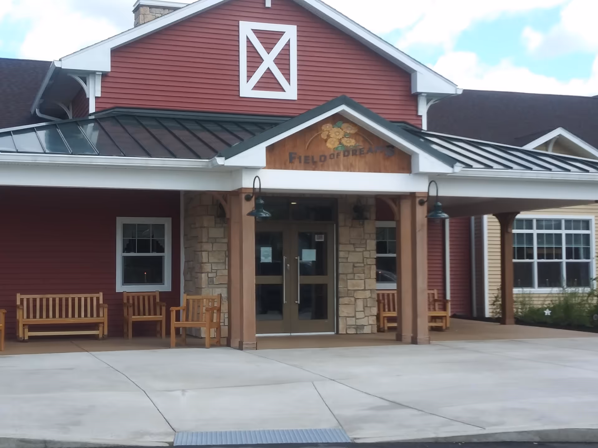 Front entrance of a building with a red exterior and stone accents. The entrance has a covered porch with wooden pillars, benches on either side, and a sign above the door that reads 'Field of Dreams' with a floral decoration.