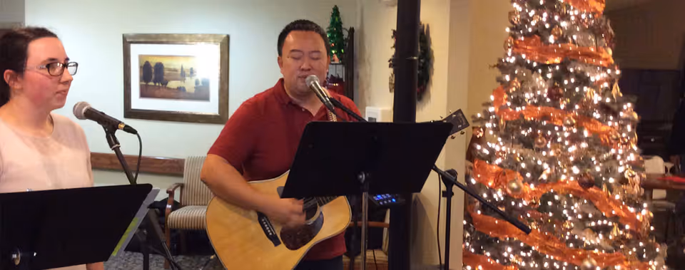 A man playing an acoustic guitar and singing into a microphone, accompanied by a woman standing next to him also singing into a microphone. They are indoors near a decorated Christmas tree with lights and orange ribbon. The background includes a framed picture on the wall and some chairs.
