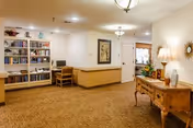 A well-lit interior common area with beige walls and carpeted floor. On the left side, there is a wooden bookshelf filled with books and a small desk with a computer. In the center, a framed artwork hangs on the wall above a wooden counter. On the right side, there is a wooden console table with a lamp, decorative items, and a mirror above it. Ceiling lights illuminate the space.
