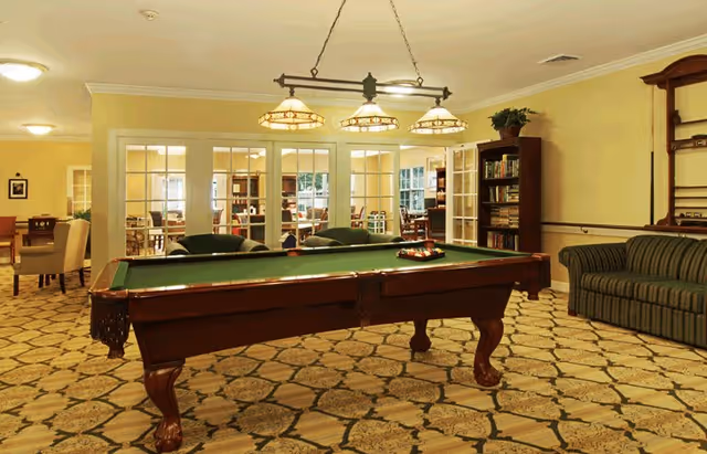 A spacious recreational room with a green felt pool table in the center, surrounded by comfortable seating including a striped couch and armchairs. The room has a patterned carpet, warm yellow walls, and a ceiling light fixture with three lamps hanging above the pool table. In the background, there are glass-paneled double doors leading to another room with more tables and chairs.