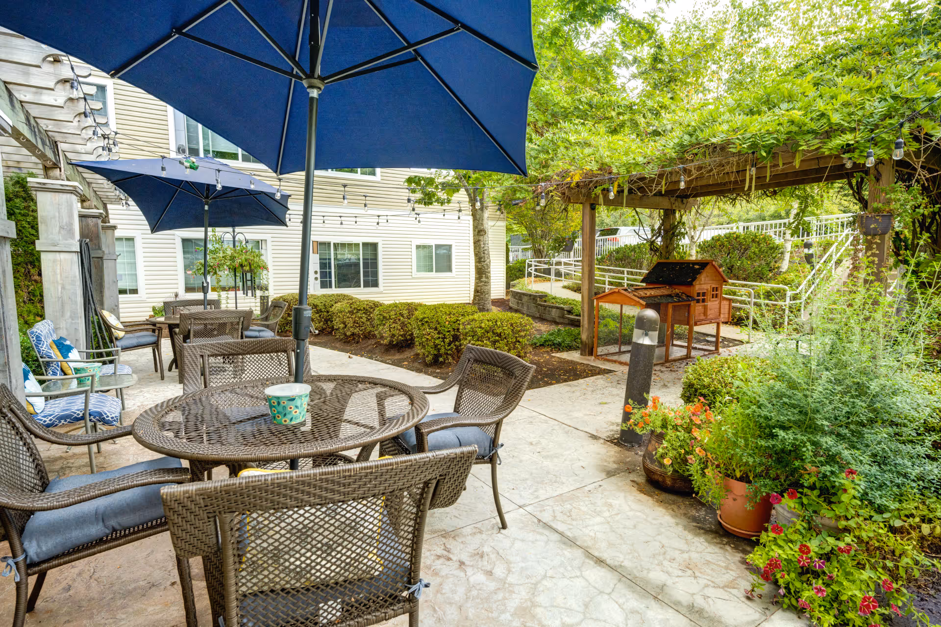 Outdoor patio area at Bay Pointe by Cogir featuring multiple round tables with wicker chairs and blue umbrellas. The patio is surrounded by greenery, potted plants, and a wooden pergola covered with vines. A small wooden structure resembling a birdhouse or decorative shed is visible near the pergola. The building exterior with windows and beige siding is in the background.