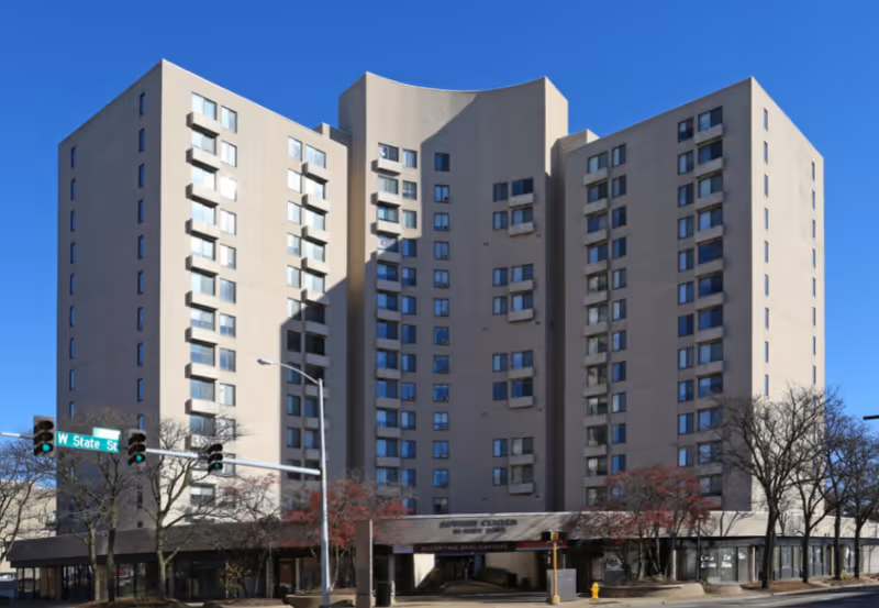 Exterior view of a multi-story beige senior living facility building named Luther Center under a clear blue sky, with leafless trees and a street intersection with traffic lights in the foreground.