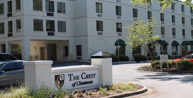 Exterior view of The Crest of Clemmons Assisted Living building with a white multi-story structure, green awnings over some windows, a parking area with cars, and a landscaped area with flowers and a sign displaying the facility's name.