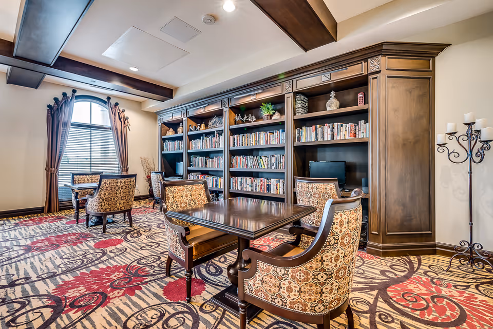 Comfortable senior living library lounge with built-in bookshelves, wooden tables, upholstered chairs, patterned carpet, and a large window with drapes.