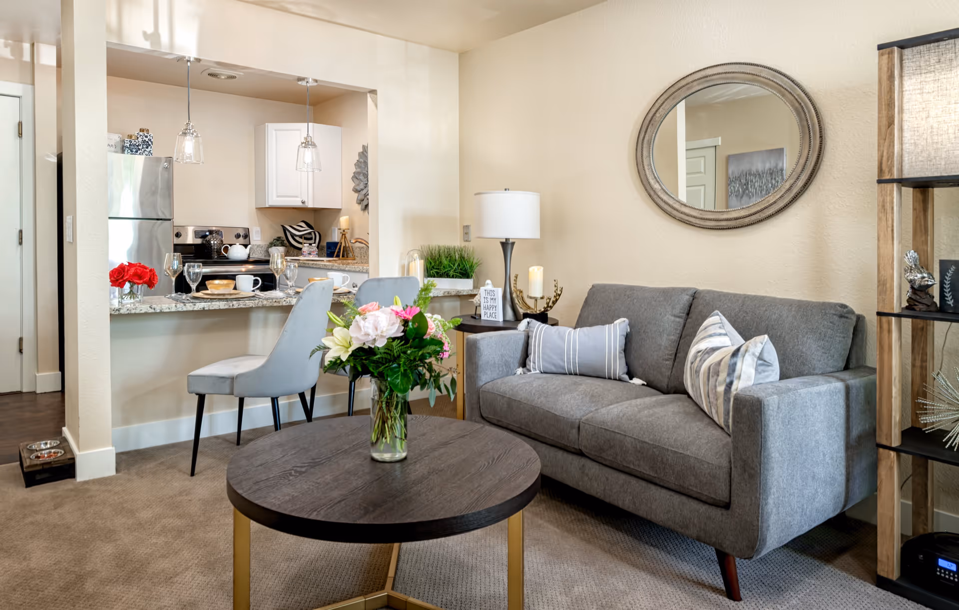A cozy living room area with a gray sofa adorned with striped pillows, a round wooden coffee table with a vase of fresh flowers, and a round mirror on the wall. Adjacent to the living room is an open kitchen with a granite countertop, two gray chairs, stainless steel appliances, and pendant lights hanging above the counter.