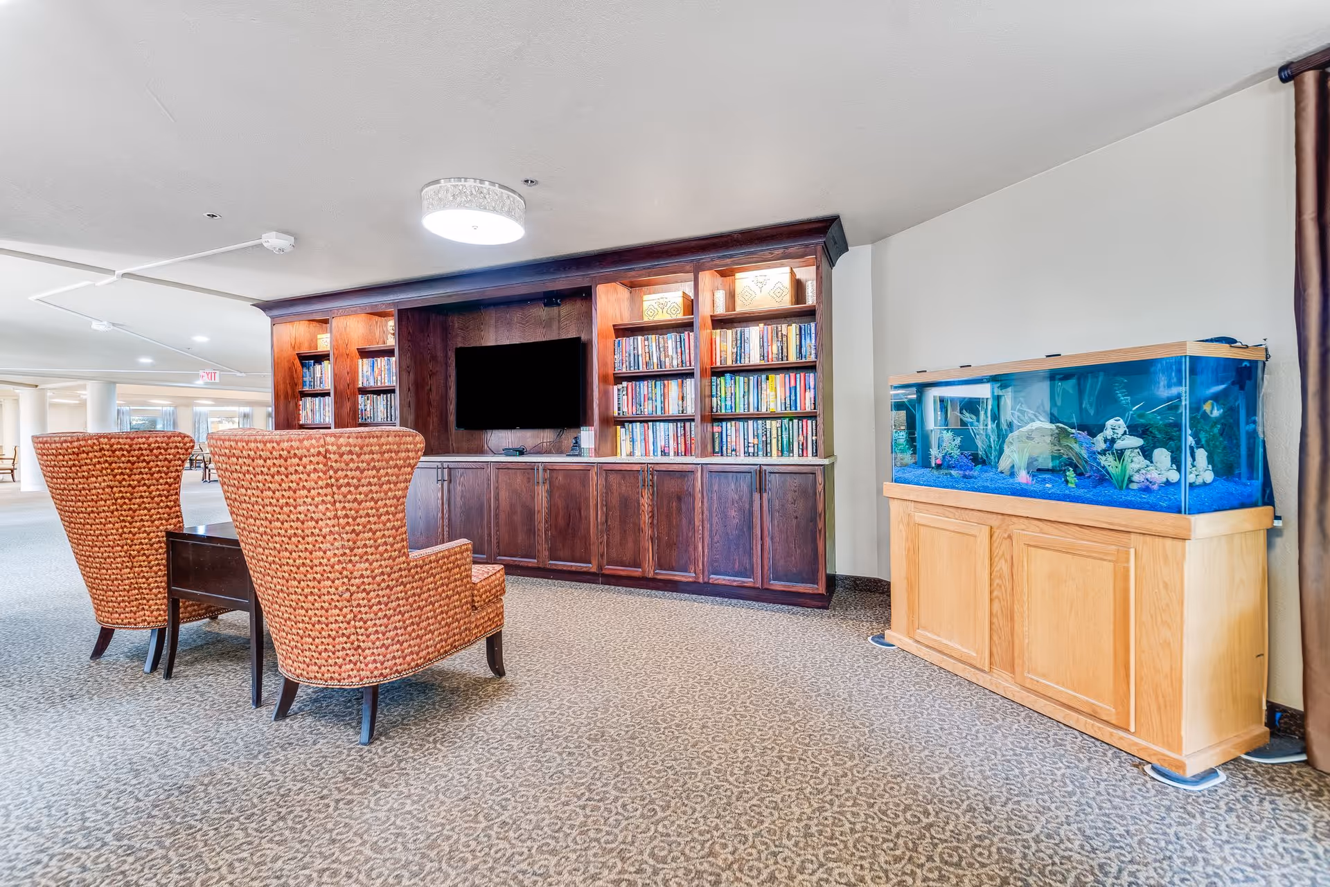 A communal seating area with two patterned armchairs facing a built-in wooden bookshelf and TV, with an aquarium on the right.