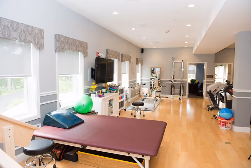 A bright rehabilitation room with light wood flooring and gray walls featuring several windows with patterned valances. The room contains a maroon therapy table with a wedge cushion, a green exercise ball, various physical therapy equipment, a wall-mounted TV, and exercise machines towards the back. There is a basket with blue and red exercise balls on the right side.