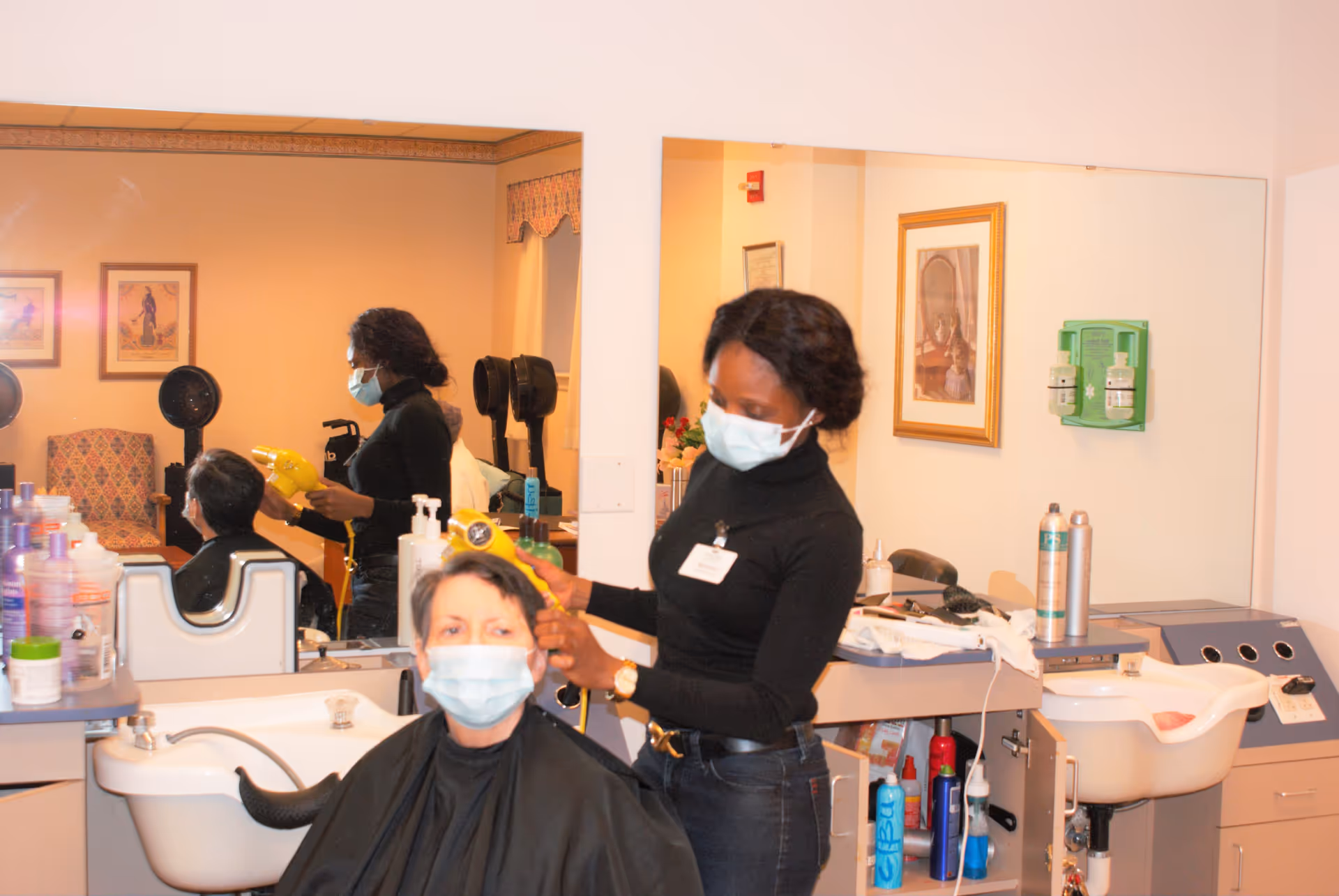 A hairstylist wearing a black top and a face mask is blow-drying the hair of a seated client who is also wearing a face mask. They are in a salon area with mirrors, hair care products, and salon equipment visible.