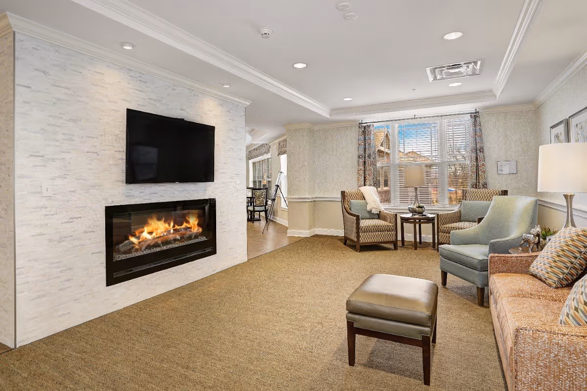 A cozy living room area with a modern electric fireplace set in a white textured wall beneath a mounted flat-screen TV. The room features a beige carpet, a brown sofa with patterned pillows, a blue armchair, two patterned armchairs with a small round table between them, and a window with patterned curtains letting in natural light. A lamp is placed on a side table next to the sofa.