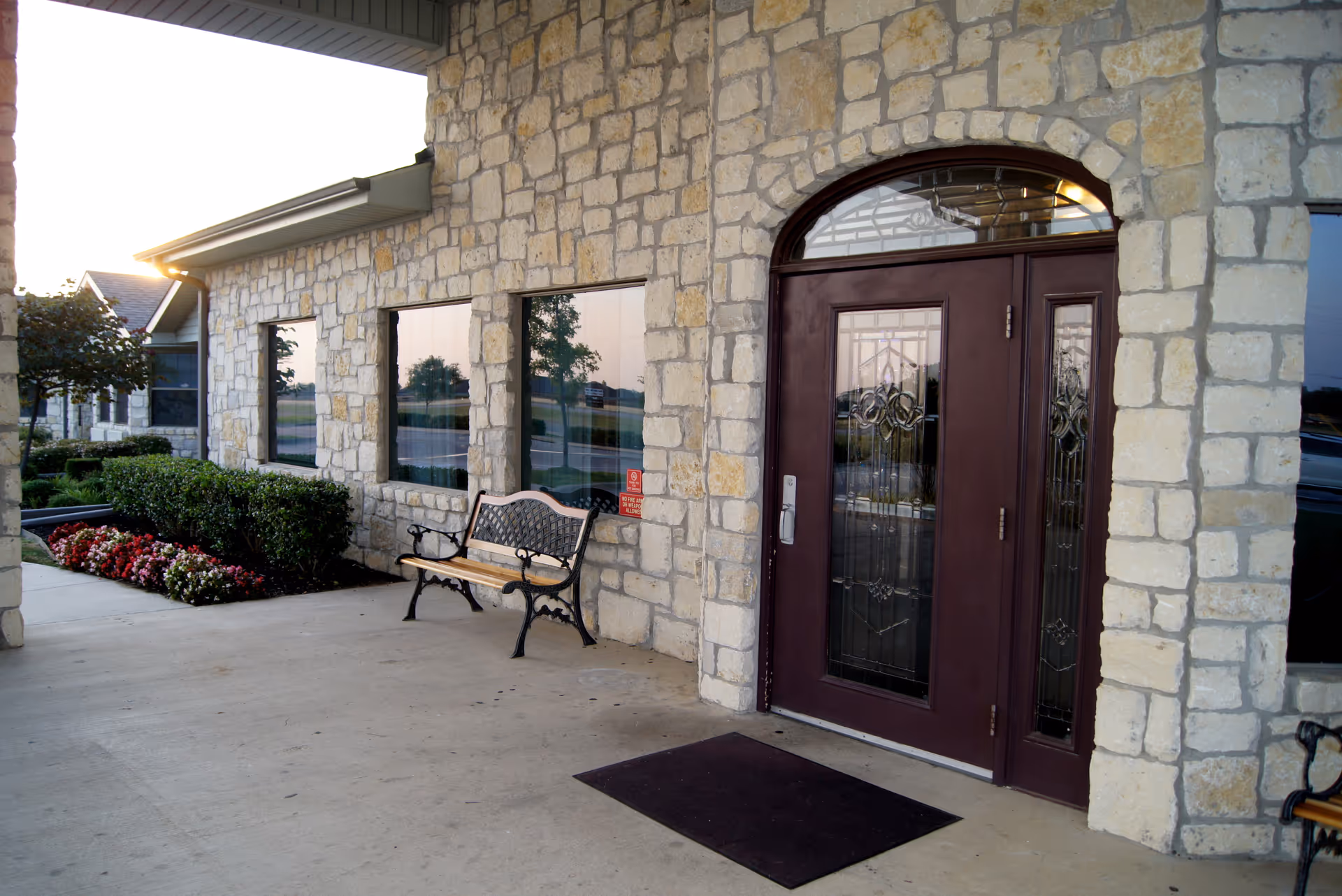 Stone exterior front entrance with decorative glass double doors, a bench, and a planted flowerbed under a covered porch.