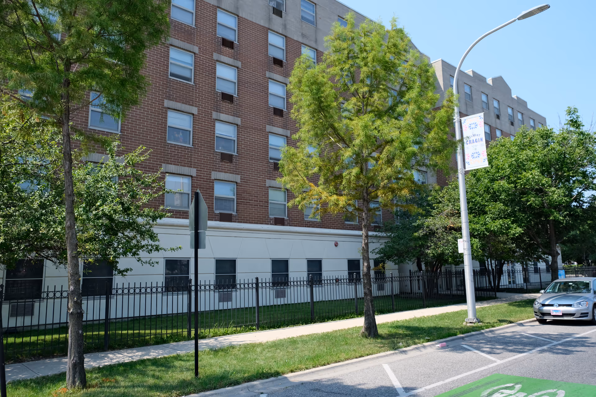 Exterior view of a multi-story brick and concrete building with several windows, surrounded by trees and a black metal fence. A sidewalk runs alongside the building, and a street with parked cars is visible. A street lamp with a banner that reads 'Belmont Cragin' is also present.