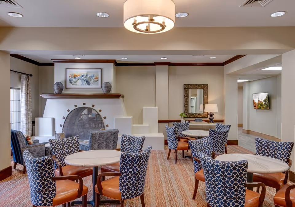 A cozy seating area in a senior living facility featuring round tables with blue patterned chairs and brown leather seats. The room has a decorative fireplace with two vases and a framed abstract painting above it. There is a large mirror on the wall with a table lamp and a small plant on a console table beneath it. The space is well-lit with ceiling lights and a central hanging light fixture, and there is a hallway visible in the background with a wall-mounted TV.