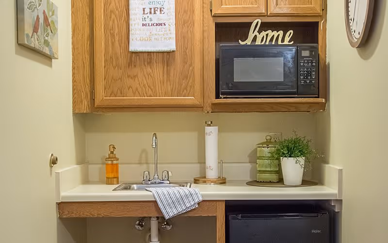 Small kitchenette area with a sink, a striped towel hanging over the sink edge, a soap dispenser, a paper towel holder, a small potted plant, a green decorative container, a microwave on a wooden shelf, and a wooden cabinet above. The wall is light-colored and there is a decorative 'home' sign on the shelf above the microwave.