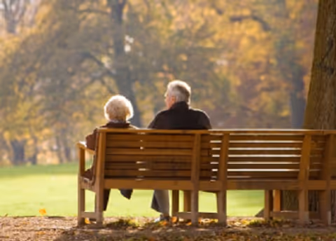 Two elderly people sitting on a wooden bench facing a sunlit park with autumn-colored trees.