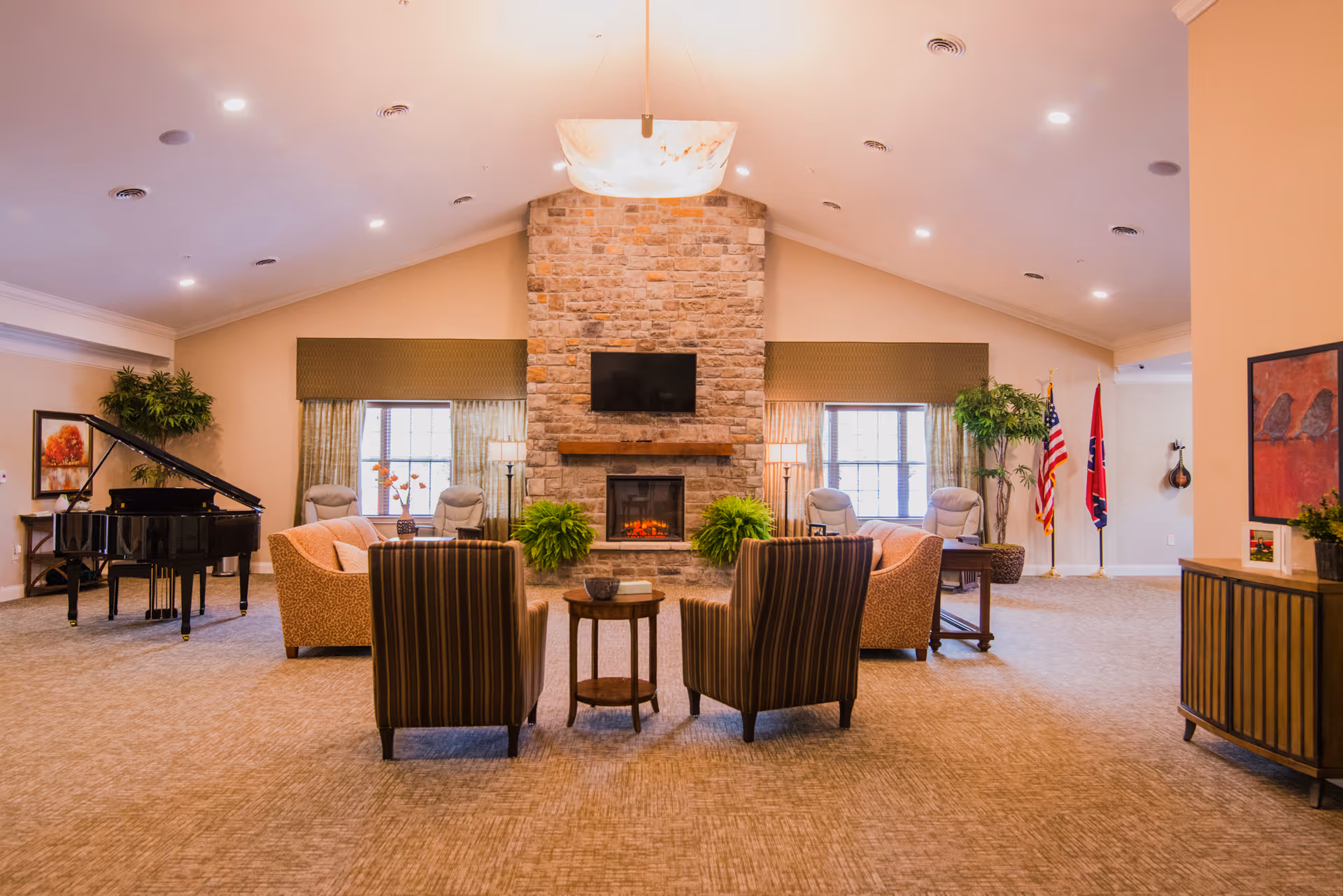 Spacious senior living common room with seating arranged around a central stone fireplace, a grand piano to the left, and flags to the right.