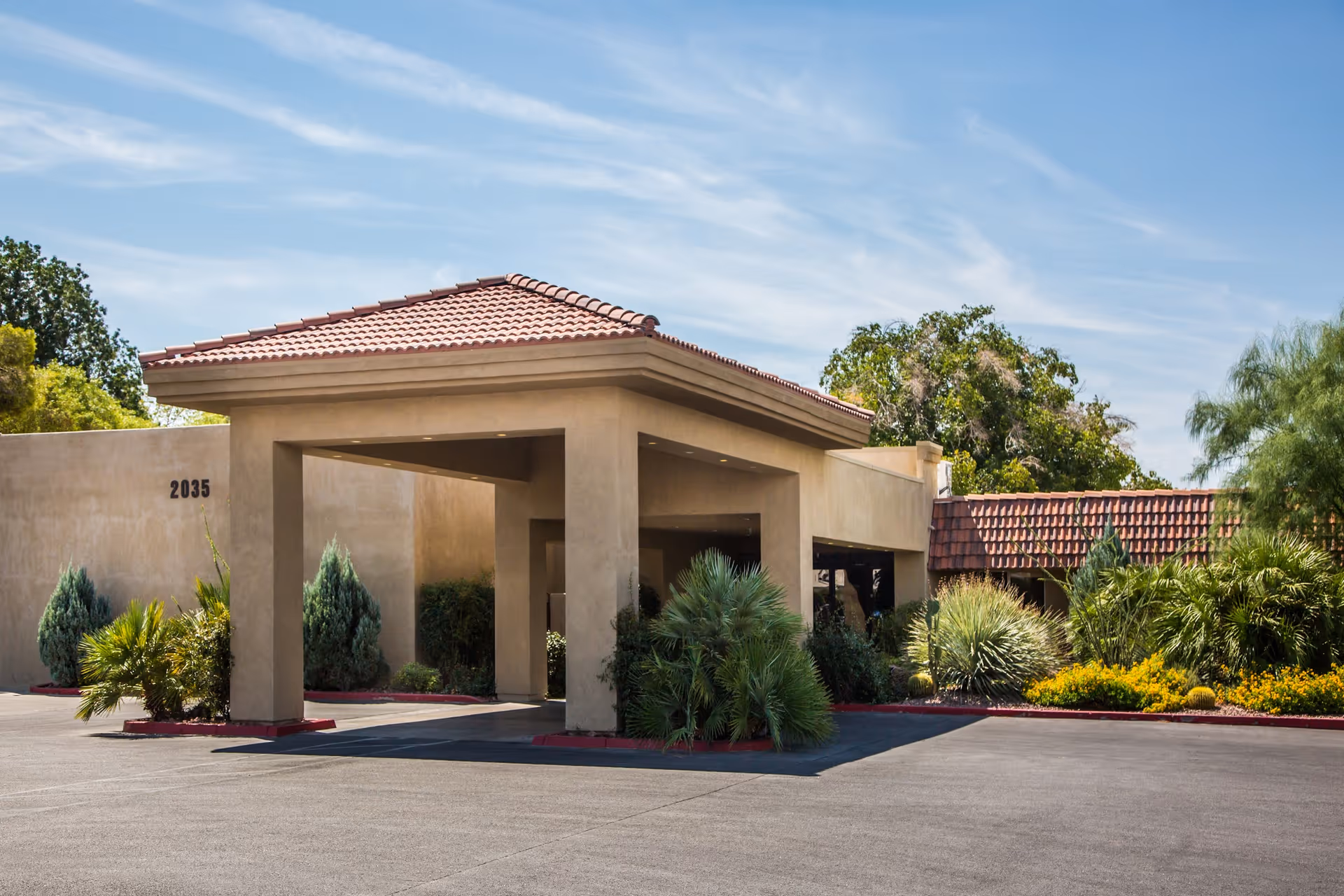 Exterior view of a building entrance with a covered driveway, surrounded by landscaped greenery and trees under a blue sky.