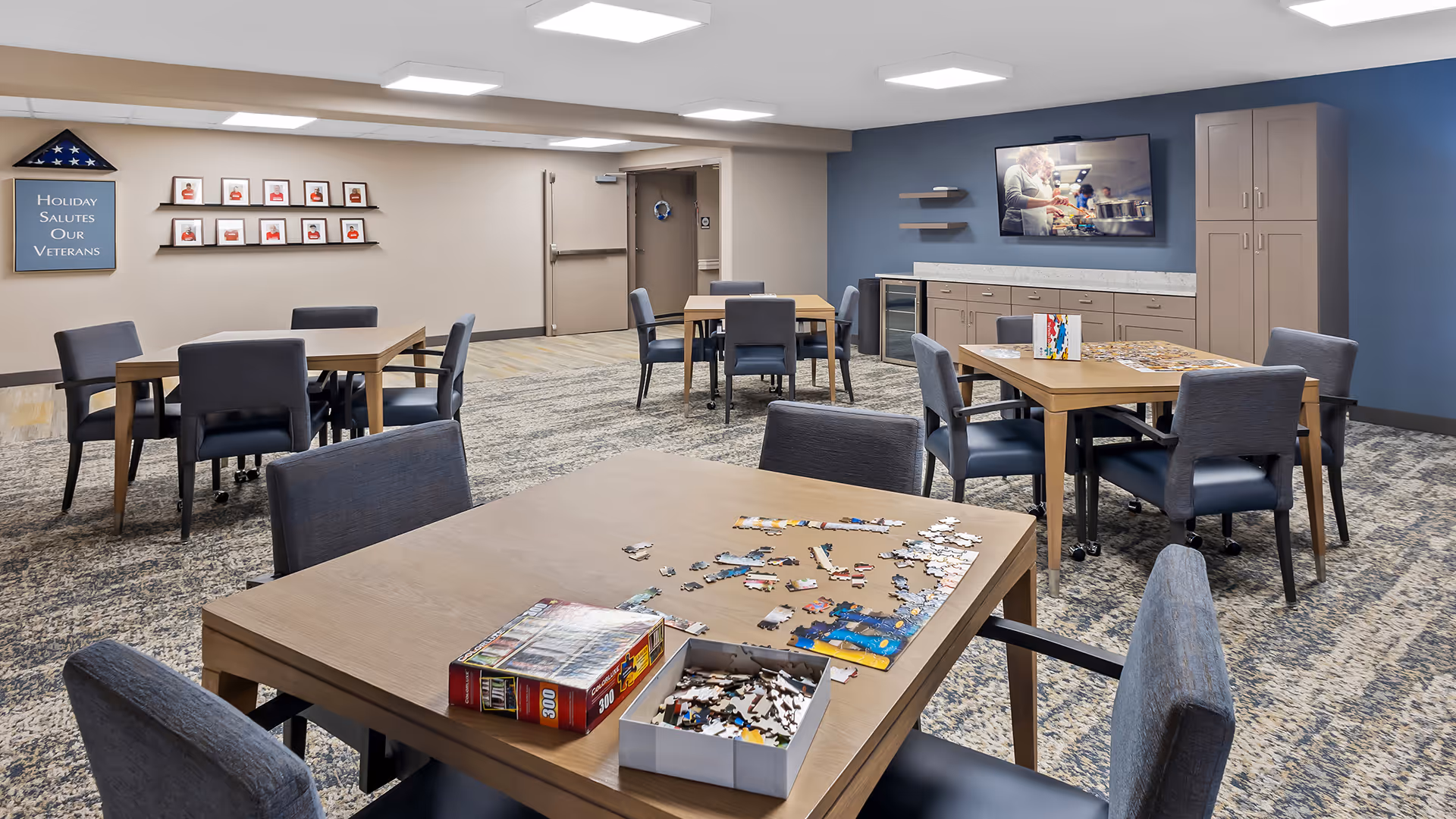 A spacious activity room with several tables and chairs arranged for group activities. On the closest table, there is a partially completed jigsaw puzzle with puzzle pieces and a box. The walls are decorated with a sign that reads 'Holiday Salutes Our Veterans' and framed pictures. A large TV is mounted on a blue accent wall above cabinets and a small refrigerator.