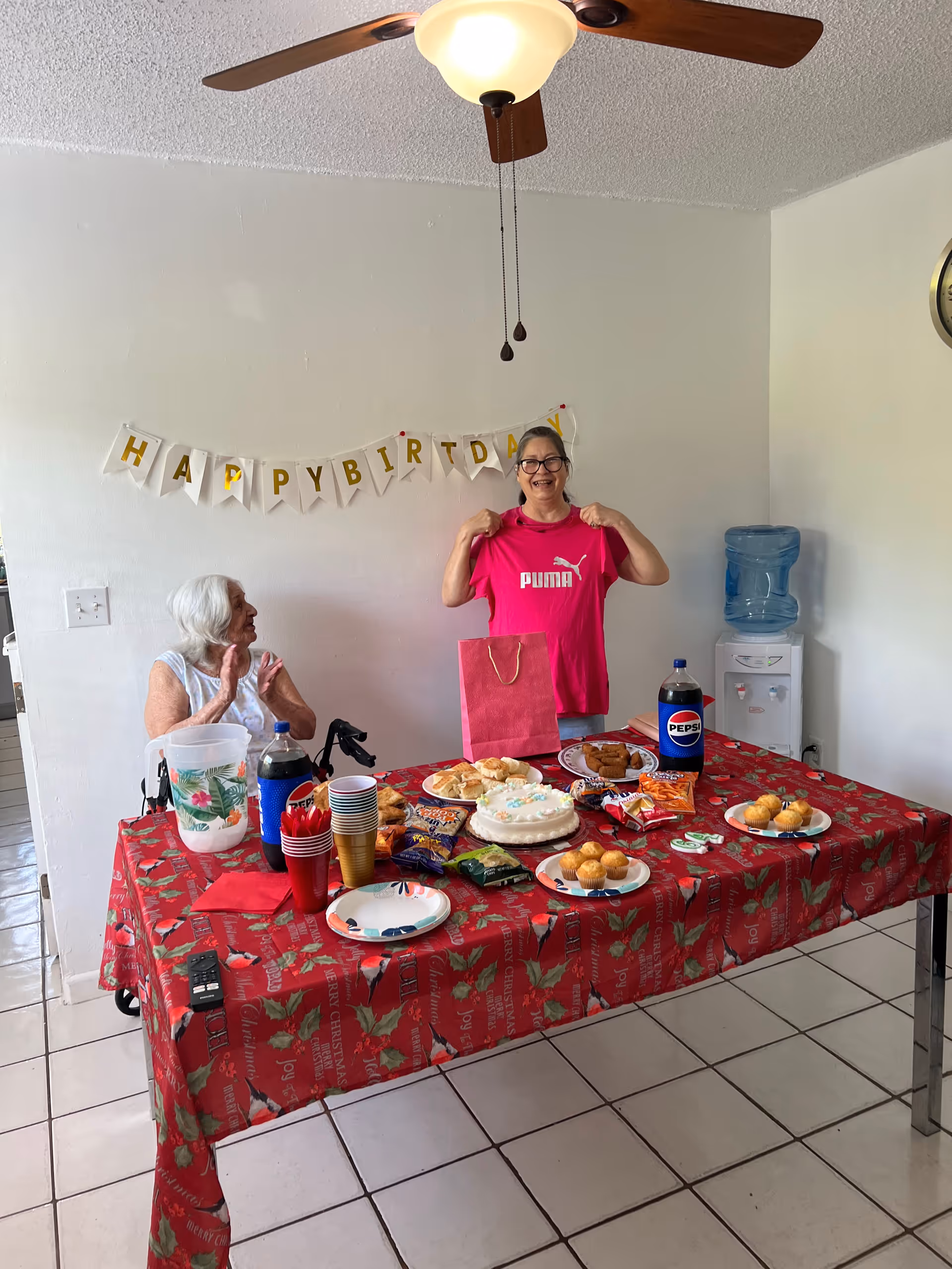 Two women celebrate at a decorated table with a birthday cake and snacks as one woman holds up a pink Puma shirt.