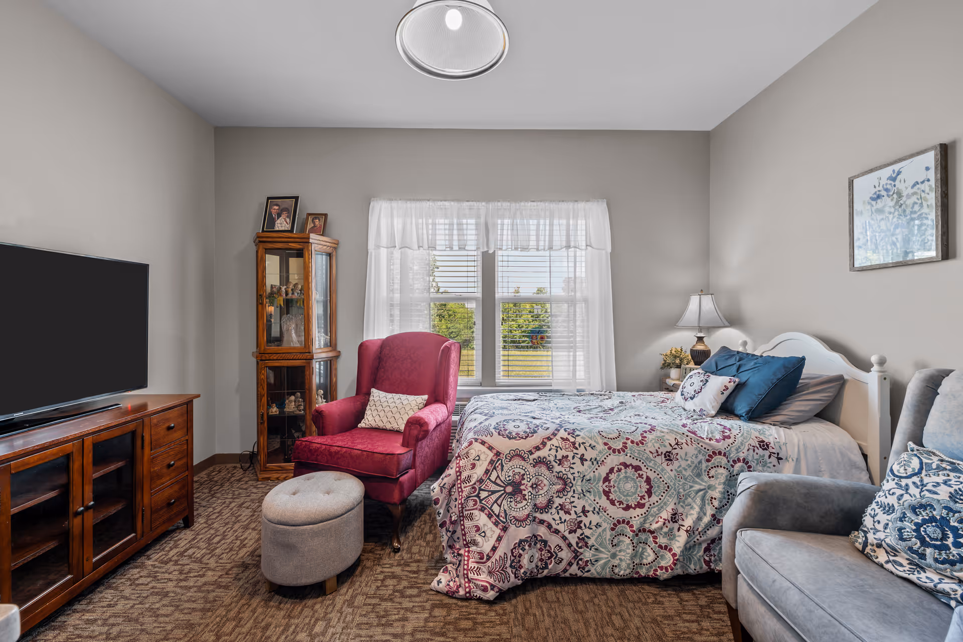 A cozy bedroom in a senior living facility featuring a bed with a colorful patterned quilt and multiple pillows, a red armchair with a small ottoman, a wooden display cabinet with framed photos on top, a TV on a wooden stand, a gray armchair with a decorative pillow, a window with white curtains letting in natural light, a bedside table with a lamp and flowers, and a framed floral artwork on the wall.