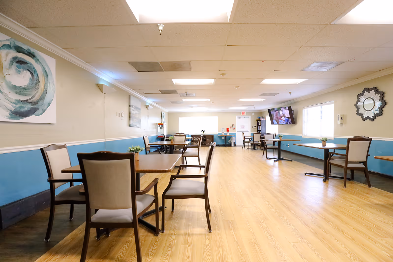A bright and spacious dining room with wooden floors, several tables and chairs arranged neatly, light blue and beige walls, abstract artwork on the wall, a wall-mounted TV, and windows allowing natural light to enter.