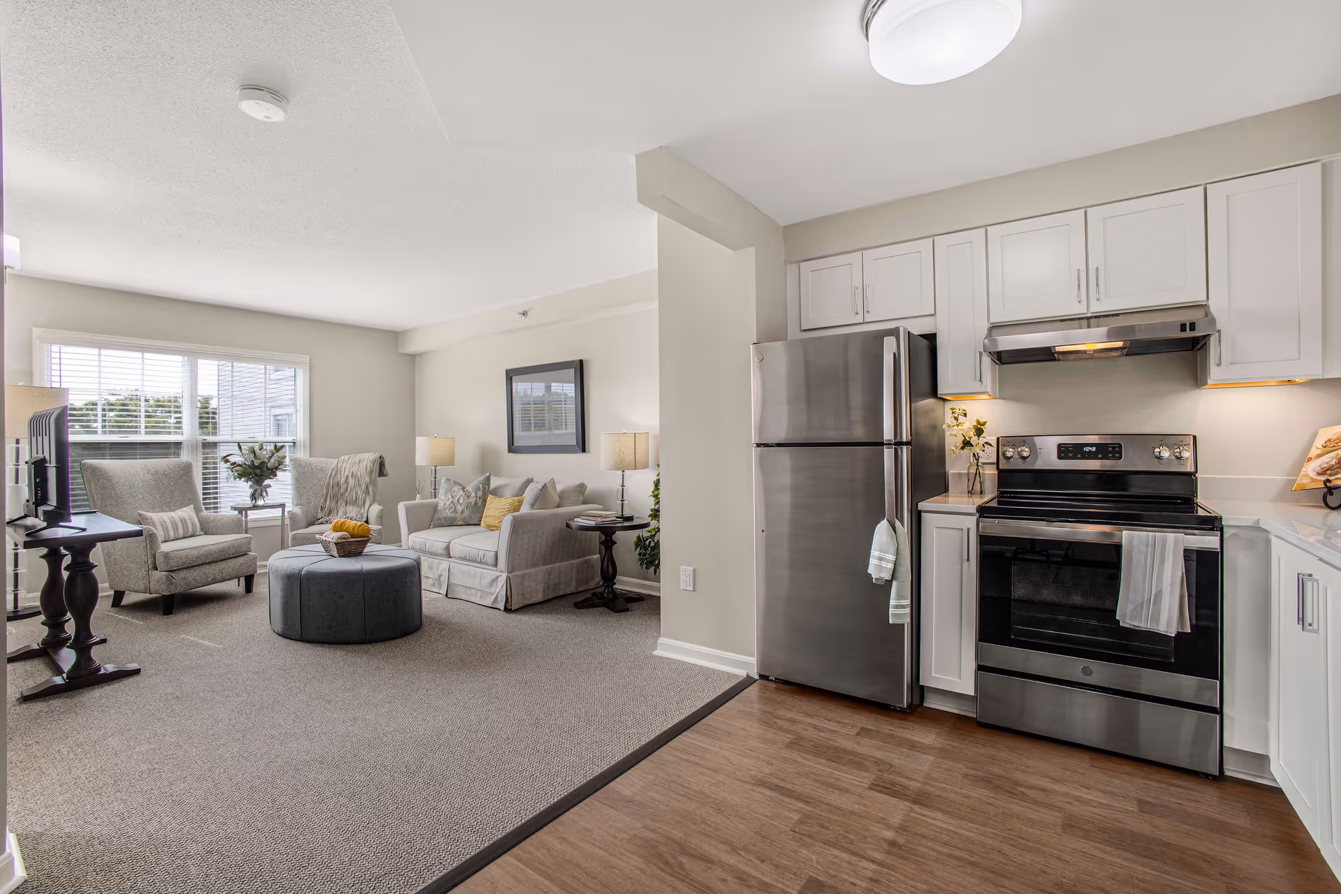 Open-plan living area and kitchen with a sofa, armchairs, round ottoman, and stainless-steel refrigerator and stove.