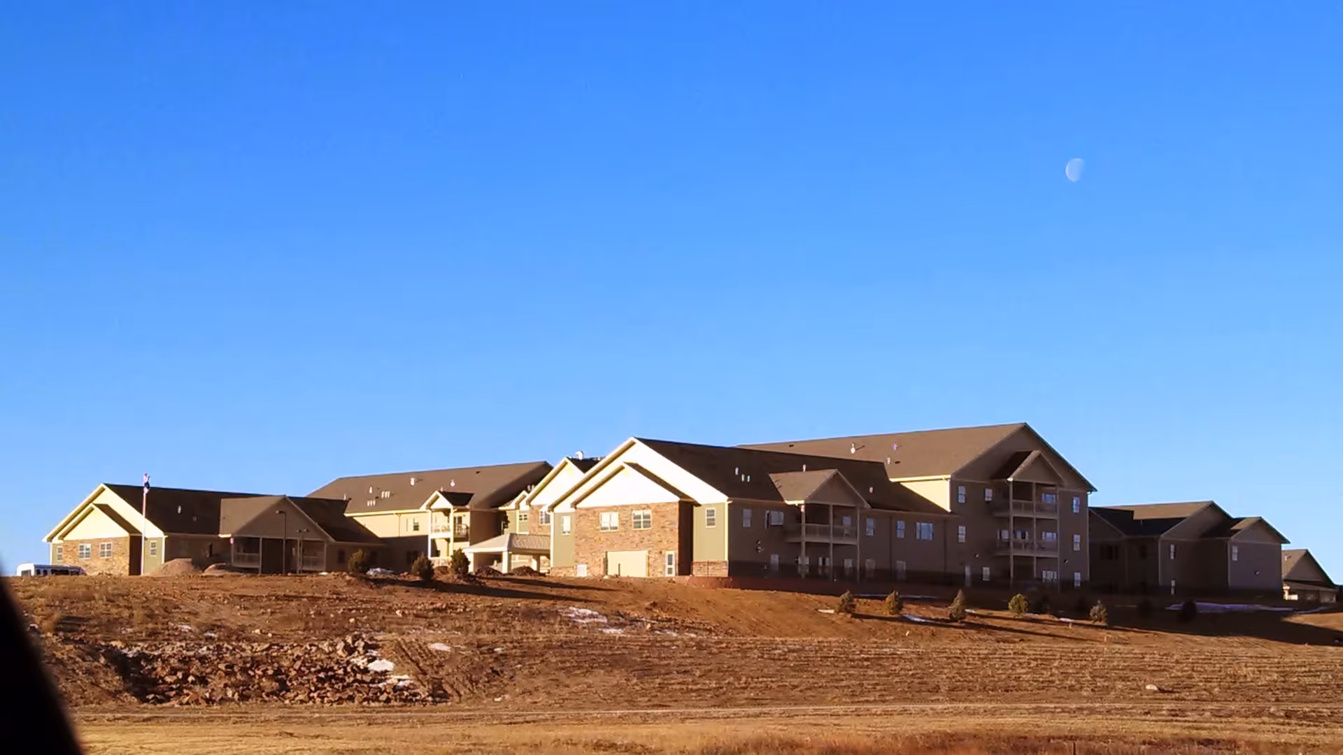 Exterior view of a large multi-building retirement community situated on a slightly elevated, barren landscape under a clear blue sky with the moon visible.