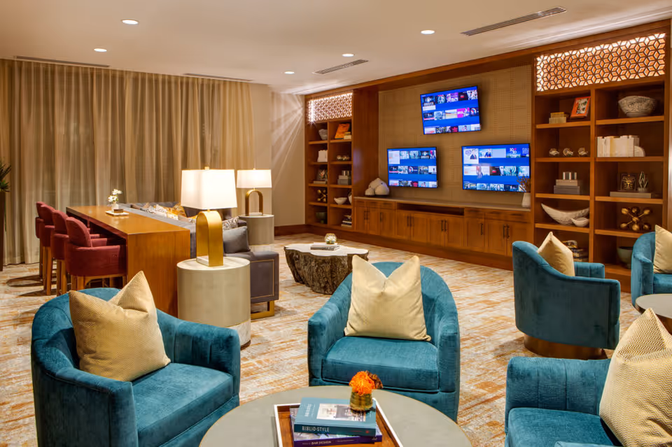 A cozy living room area with teal armchairs and beige pillows arranged around a round coffee table with books and a small flower vase. Behind the seating area is a wooden console table with red chairs and two modern lamps. The back wall features built-in wooden shelves with decorative items and three mounted flat-screen TVs. The room has warm lighting and beige curtains covering the windows.
