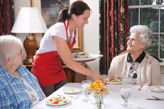 A server in a red apron sets plates of salad for two elderly women seated at a dining table with glassware and a floral centerpiece.