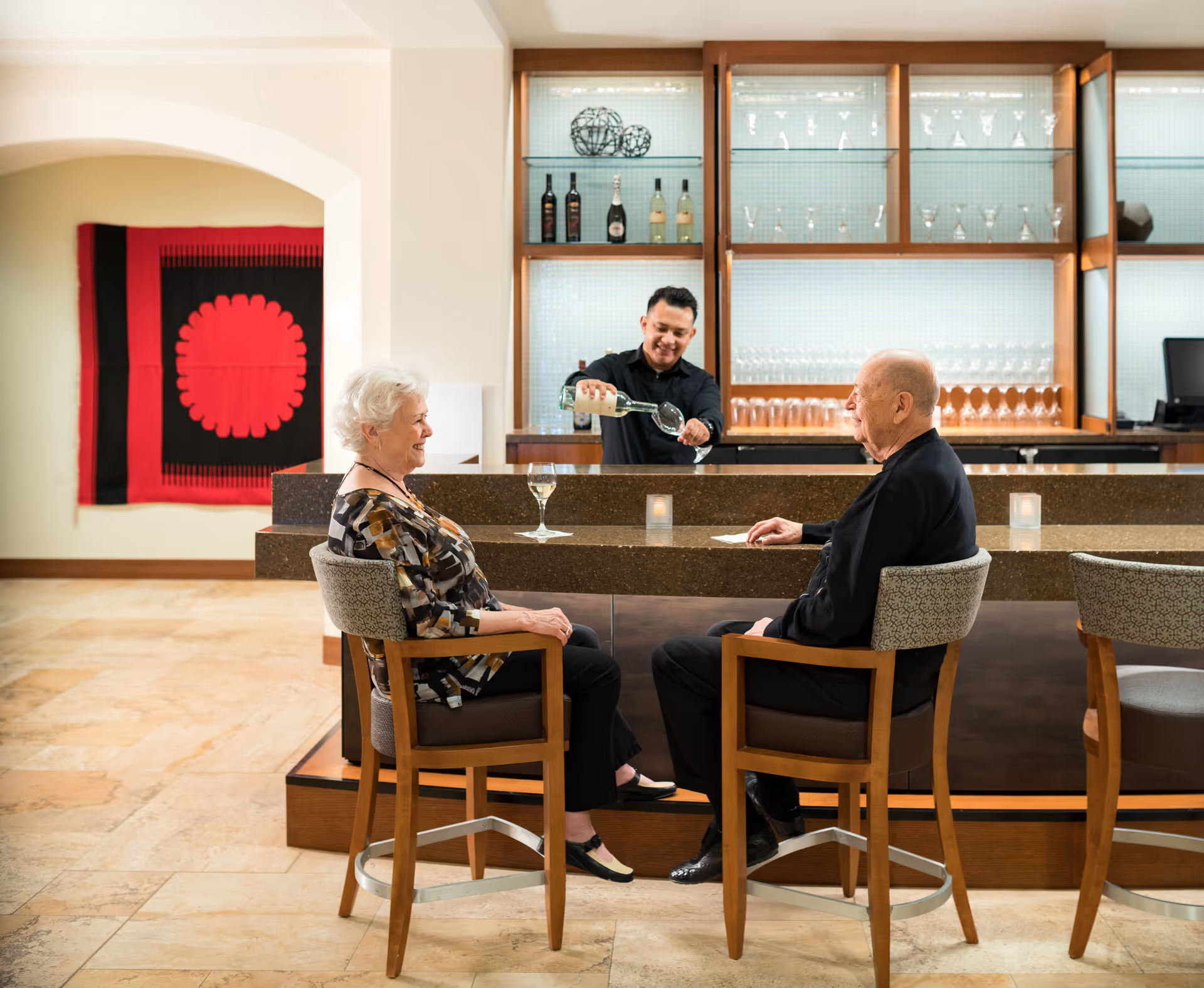 An elderly woman and man sit on bar stools at a counter while a bartender pours a glass of white wine for them. Behind the bartender, shelves display bottles and glassware. The setting appears to be a comfortable indoor bar area with warm lighting and a decorative red and black wall hanging in the background.
