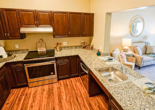 Open kitchen with granite countertops, dark wood cabinets, stainless steel stove and sink overlooking a nearby seating area.