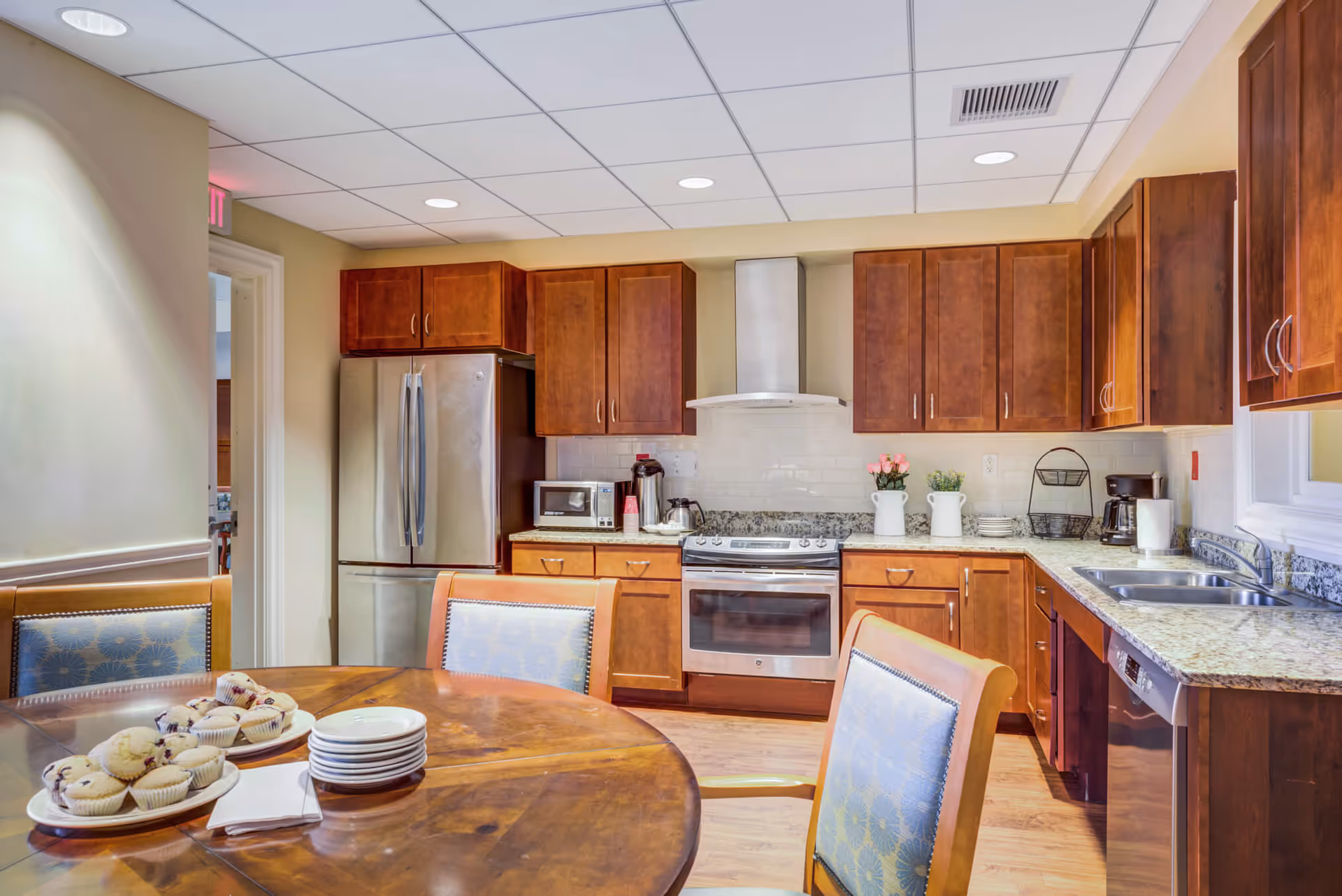 A bright kitchen area with wooden cabinets, stainless steel refrigerator, oven, and dishwasher. A round wooden table with blue cushioned chairs is in the foreground, with plates and muffins on the table. The kitchen has a granite countertop, a double sink, and a coffee maker. There are decorative flowers on the counter and recessed lighting in the ceiling.