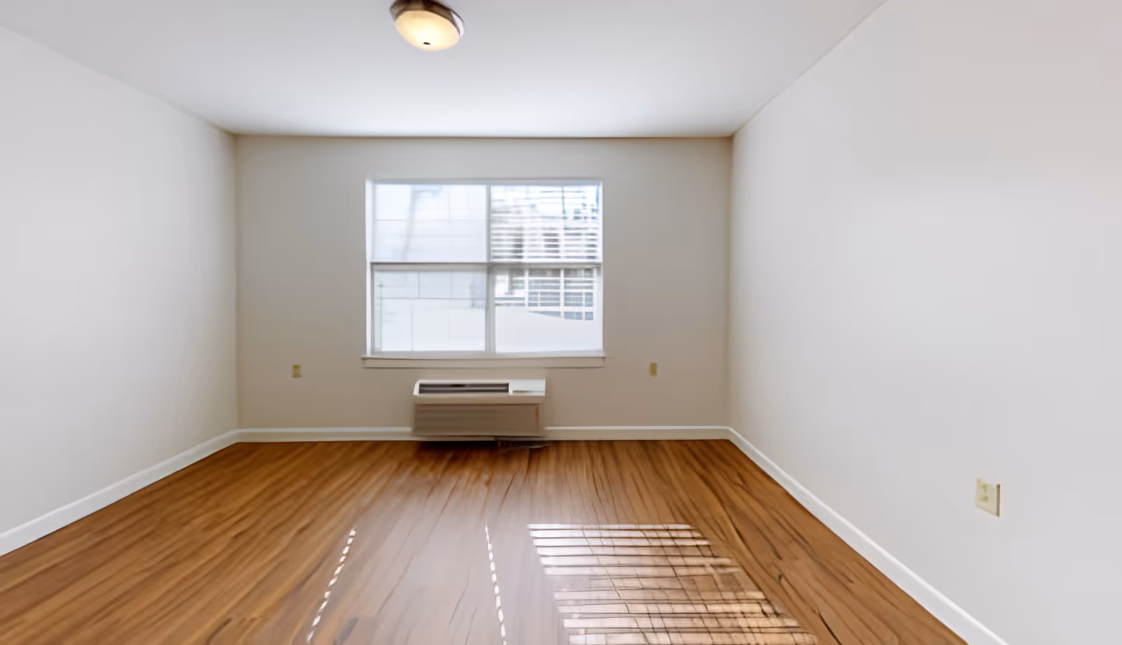 Empty rectangular room with wood floors, white walls, a window with blinds and a wall-mounted HVAC unit beneath it.