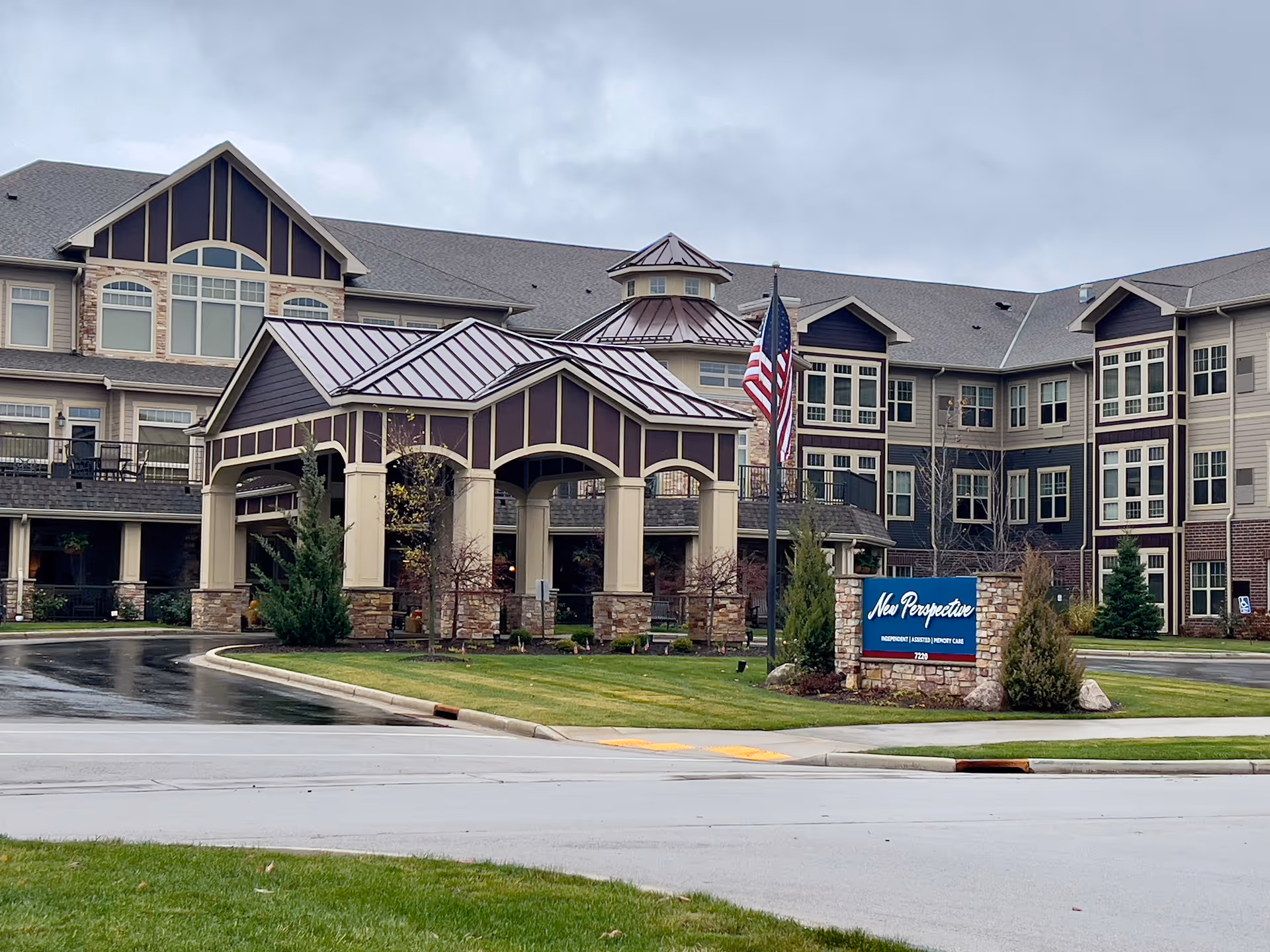 Exterior view of New Perspective Senior Living | Franklin building on a cloudy day, featuring a covered entrance with stone pillars, an American flag, and a sign displaying the facility's name and services.