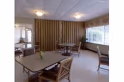 A dining area with several tables and chairs arranged neatly. The room has large windows with curtains allowing natural light to enter. There are small flower vases placed on the tables. The walls are light-colored and there is a wooden partition in the background.