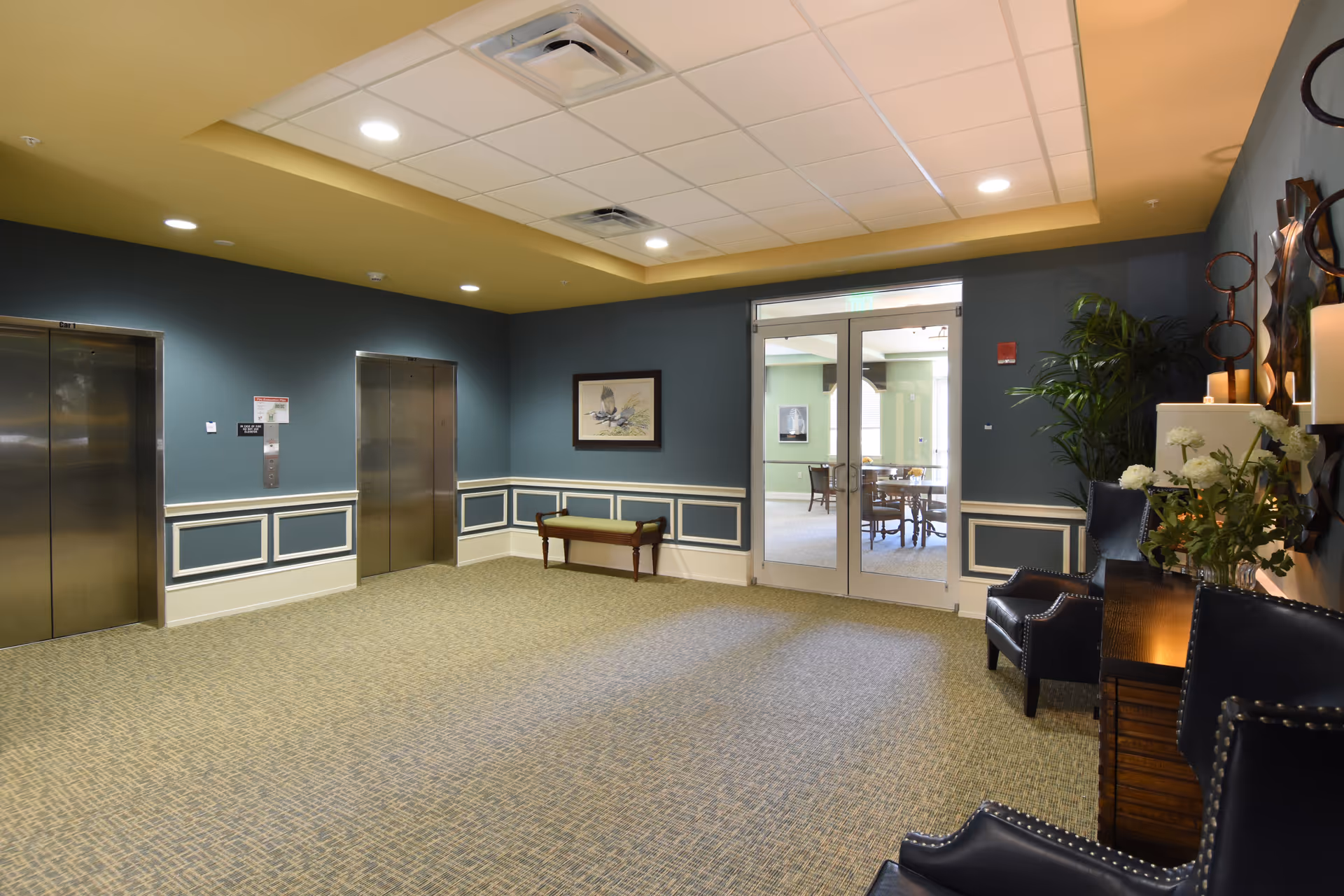 Well-lit interior lobby with two elevators on the left, seating and decorative furniture on the right, and double doors leading to a dining area.
