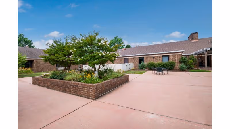 Outdoor courtyard area with a raised brick planter containing green trees and flowering plants. Surrounding the planter is a spacious paved area with a small table and chairs. The courtyard is enclosed by single-story brick buildings under a blue sky with some clouds.
