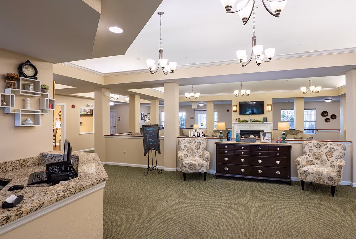 Interior view of a senior living facility lobby area with a granite reception desk on the left, two patterned armchairs, a dark wooden dresser with decorative items, a fireplace with a TV mounted above it, and multiple ceiling light fixtures illuminating the space.
