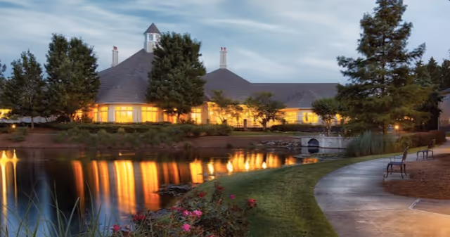 Evening view of a large building with warm interior lights reflecting on a pond in front. The building has a steep roof with a small cupola on top. There is a curved walkway along the pond with benches and landscaped greenery including trees and flowers.